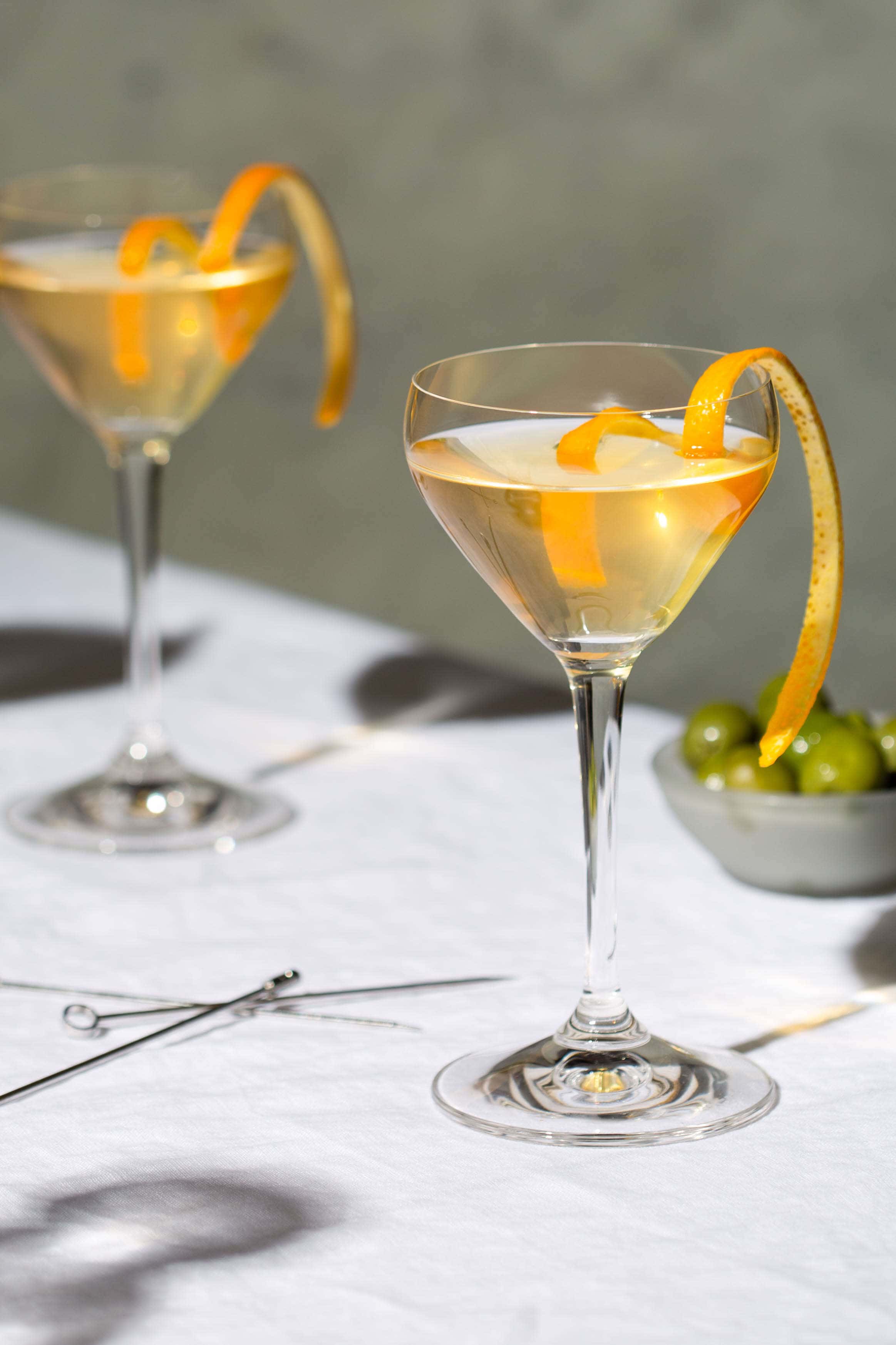 cocktail in a nick nora glass with a garnish of orange and olives in a bowl in the background