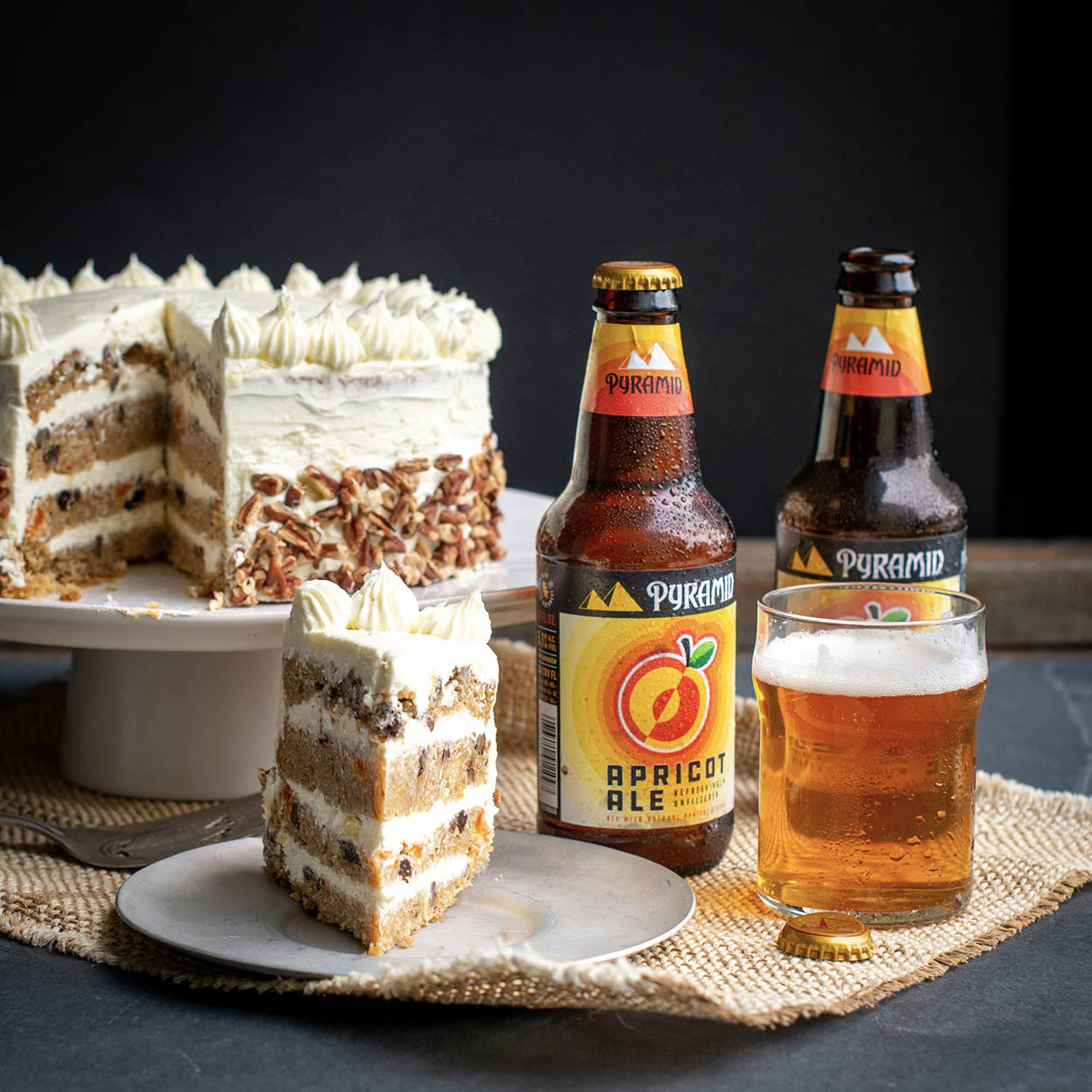 Best carrot cake recipe with pineapple frosting and beer ingredients A slice of homemade carrot cake served with a glass of apricot ale, the dessert served in front of the whole carrot cake, tastefully decorated with a cream cheese pineapple frosting, two bottles of pyramid apricot ale linger to the side of the photo paying homage to the cake's signature ingredient - Pyramid Apricot Ale