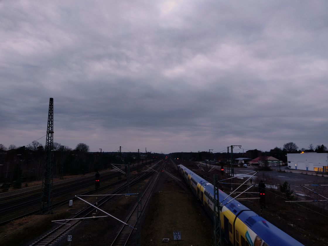 Clouded evening sky above dimly lit rail tracks that converge at the horizon. A passenger train on the right with cars painted blue, yellow, and white.