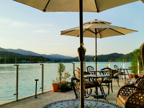 Scenic outdoor cafe deck with cream umbrellas over ornate tables and chairs in front of mountain lake view Google Meet background