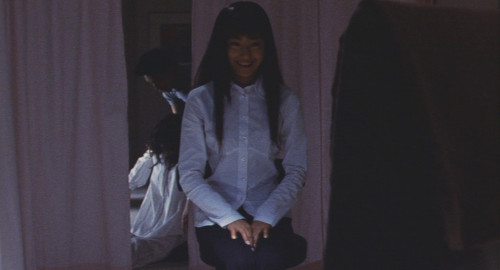 A screenshot of a young woman Tomie (played by Miho Kanno), sitting on a chair looking menacingly at another girl off-screen from the movie 'Tomie'.