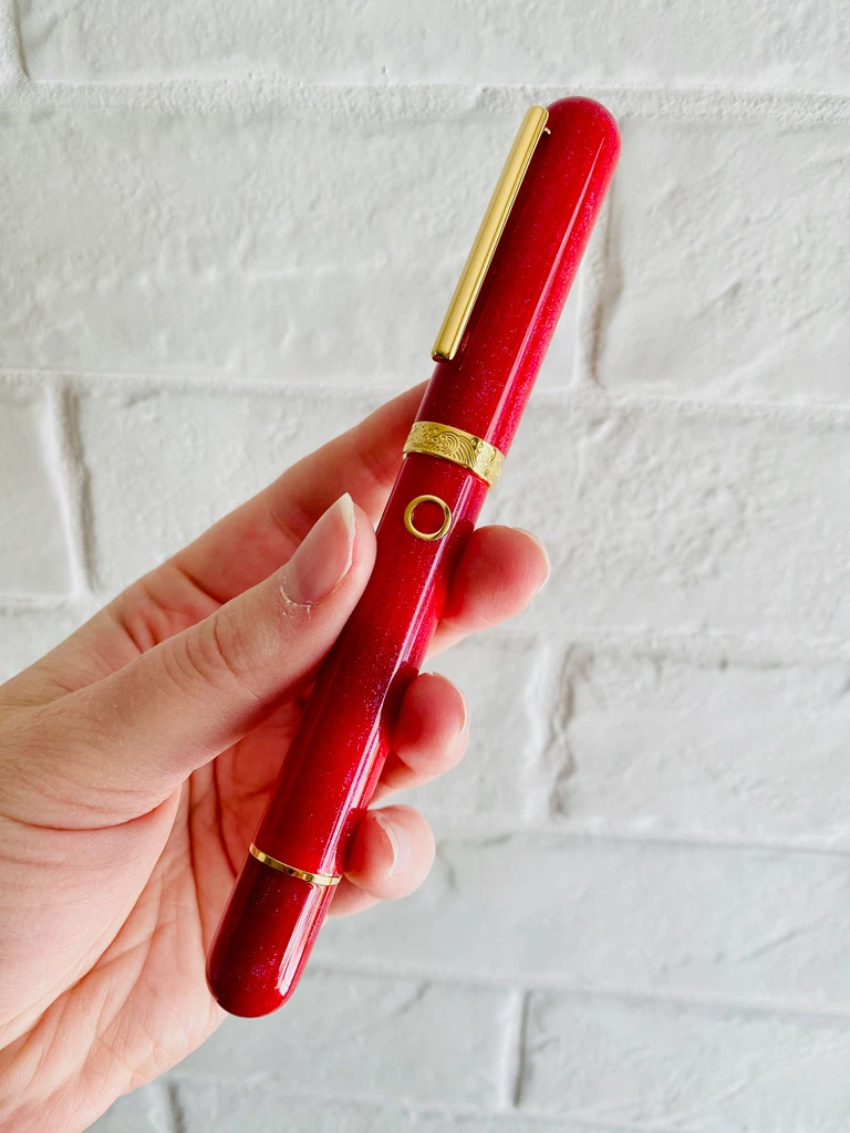 A red, sparkly fountain pen with gold trip on the cap and clip. There is a circular ink window that looks like a port hole on a ship.