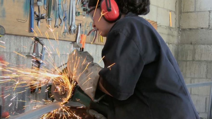 a woman wearing protective gear uses an angle grinder to cut a piece of metal (photo credit: actuality media)
