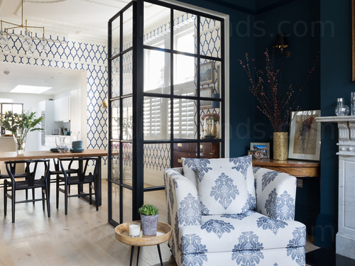 Living room corner featuring arm chair with white and blue pattern and interior window door with kitchen beyond  Skype background
