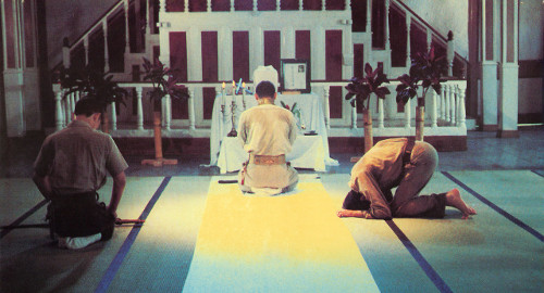 A screenshot from the movie 'Merry Christmas, Mr. Lawrence' of three men on their knees praying to an alter in a Japanese ceremonial hall shown from behind.