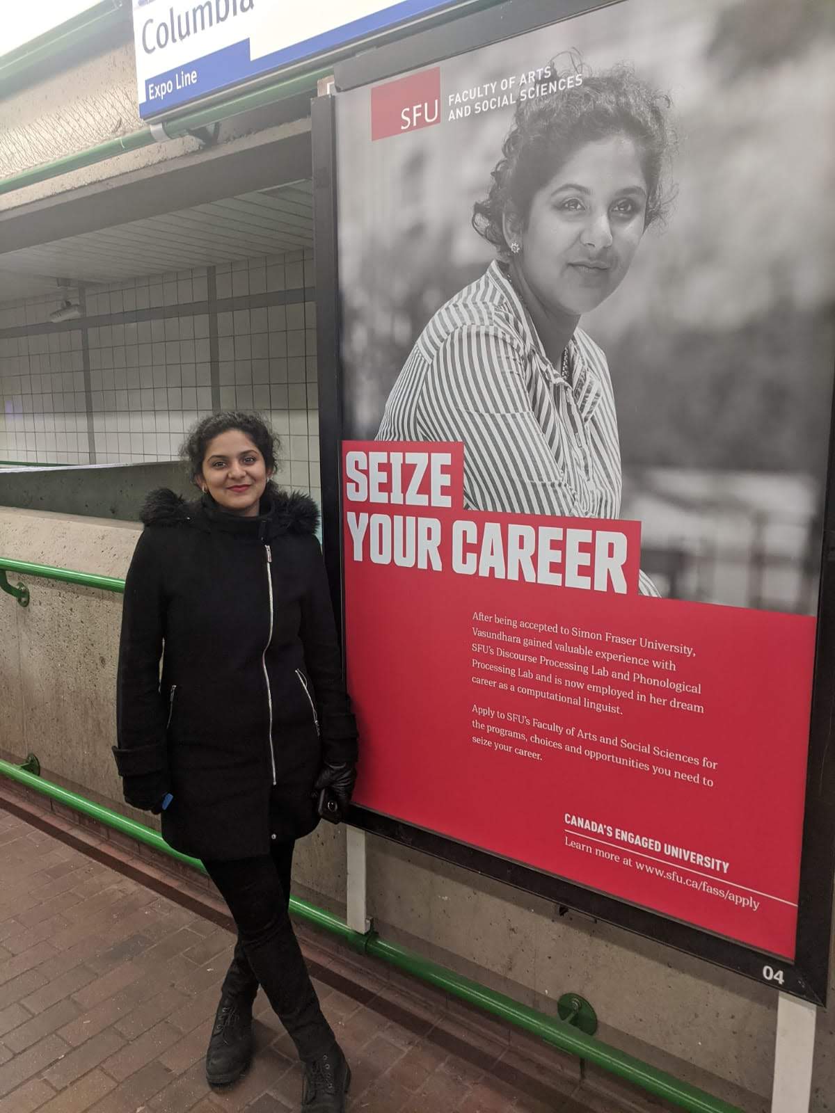 Vasundhara standing in a SkyTrain station beside a large advertisement for Simon Fraser University's Faculty of Arts and Social Sciences. The advert has a black-and-white photograph of xem in it, and the words 'Seize Your Career' in big, bold text, followed by two paragraphs of small text talking about xyr career trajectory and why readers should apply to the Faculty of Arts and Social Sciences.