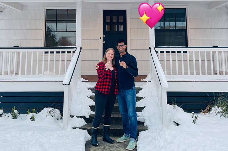 My boyfriend and I holding up keys in front of a cream-colored farm-style house with black and white trim. There is heavy snow on the roof and small lawn in front of the house.