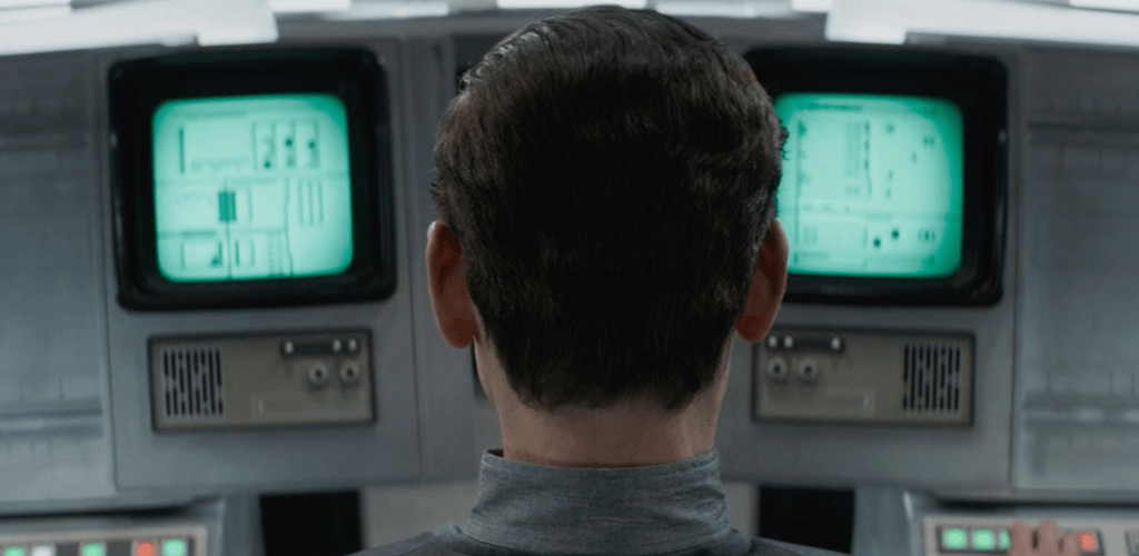 A screenshot from the series Andor, showing the back of a mans head with black hair, in front of two monochromatic-green computer displays.