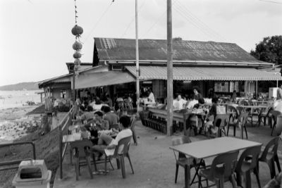 A black and white photo of Ponggol (Hock Kee) Seafood Restaurant, showing an outdoor dining area facing the sea. The coffee shop-style tables closest to the seafront are occupied by diners.