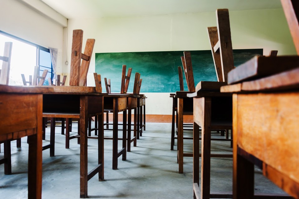 An empty classroom with old wooden desks, chairs placed upside down on desktops, and an old-fashioned chalkboard.
