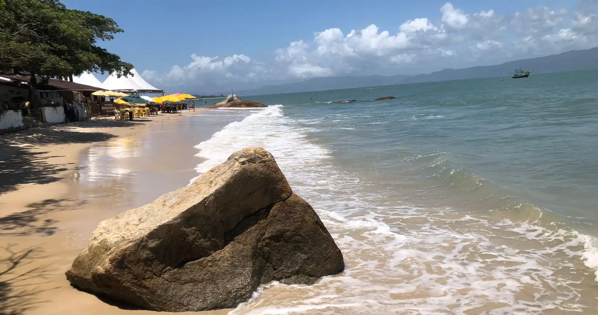 Grande pedra na area com onda batendo levemente e faixa de area atrás mostrando um bar ao fundo com alguns guarda-sóis