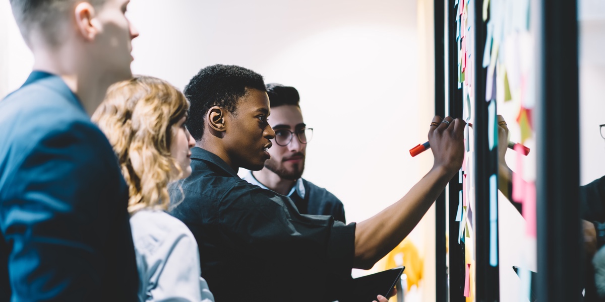 What Is Inclusive User Research? Group of UX designers gathered around a whiteboard with sticky notes, watching one designer sketch their ideas out