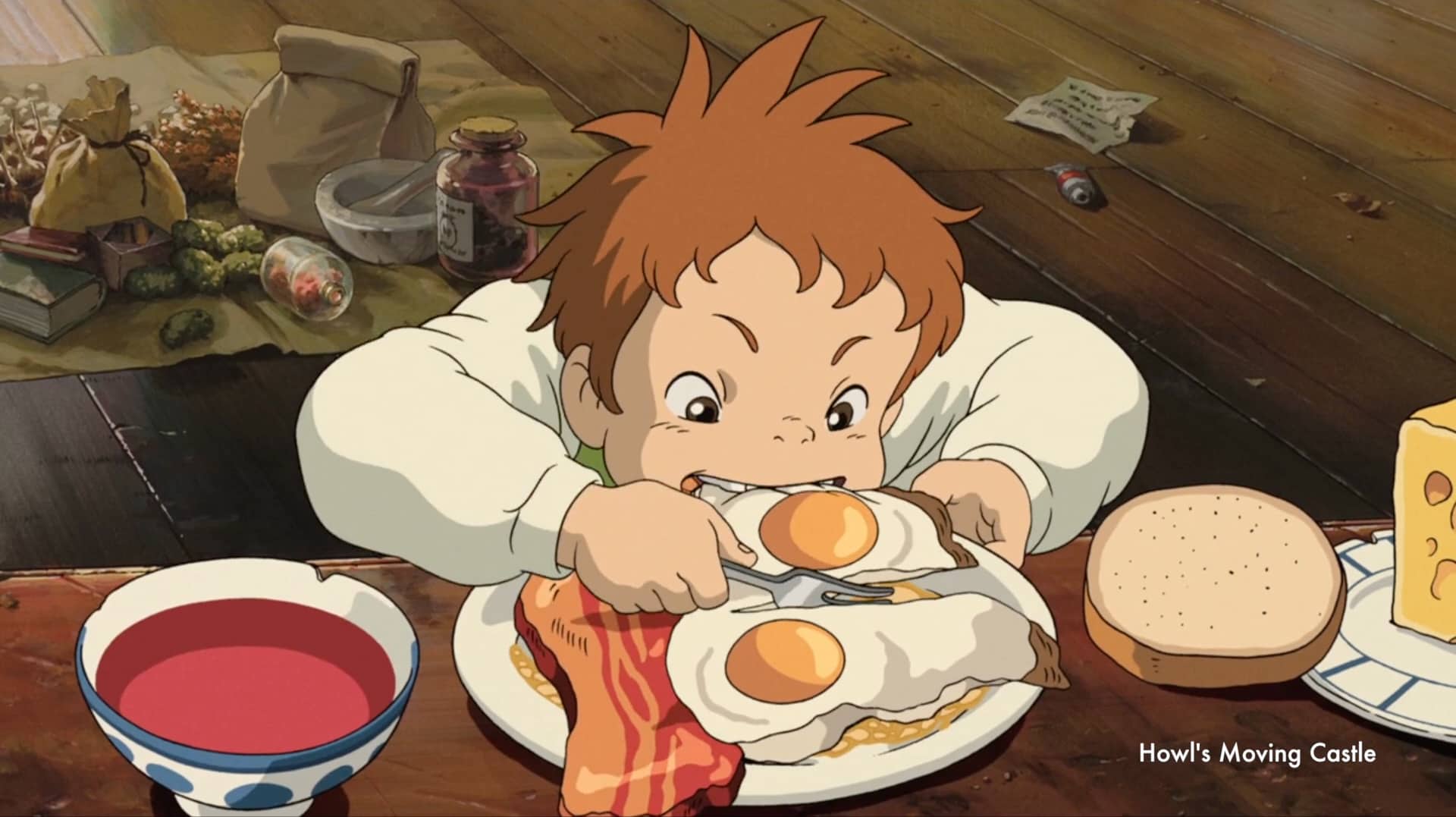 Single frame from the anime Howl's Moving Castle. The table is set with food: soup, bread, cheese, egg, and meat. A boy gobbles the eggs with enthusiasm.