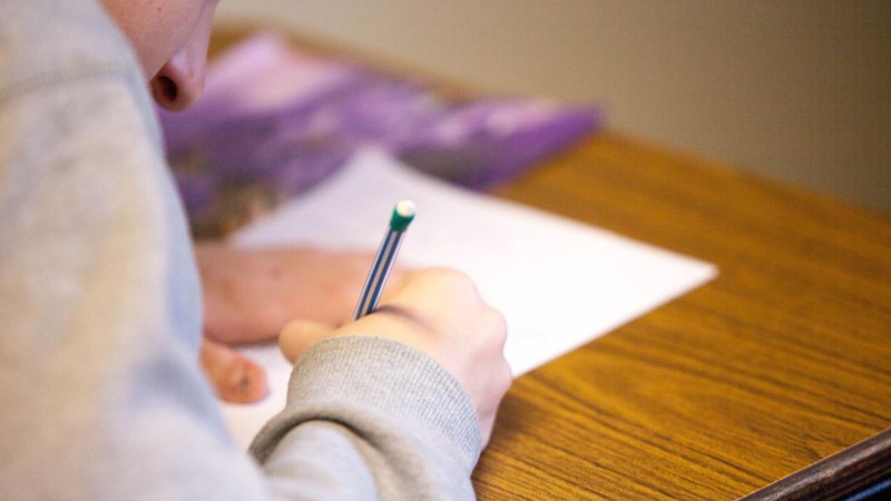 A student writes with a pencil on a white piece of paper.