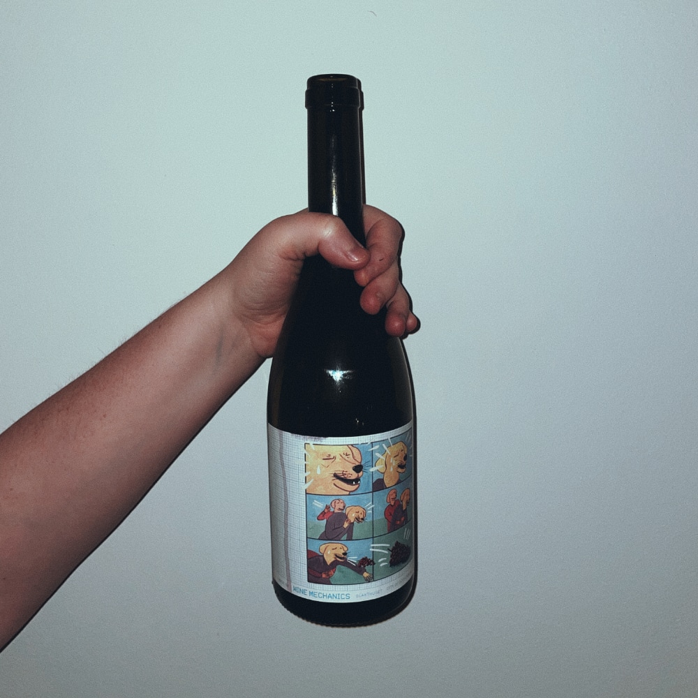 Someone holding a wine bottle against a white wall. The label is illustrated, depicting two anthropomorphized dogs. One performing the Heinrich maneuver on the other, who is coughing up a branch of grapes.