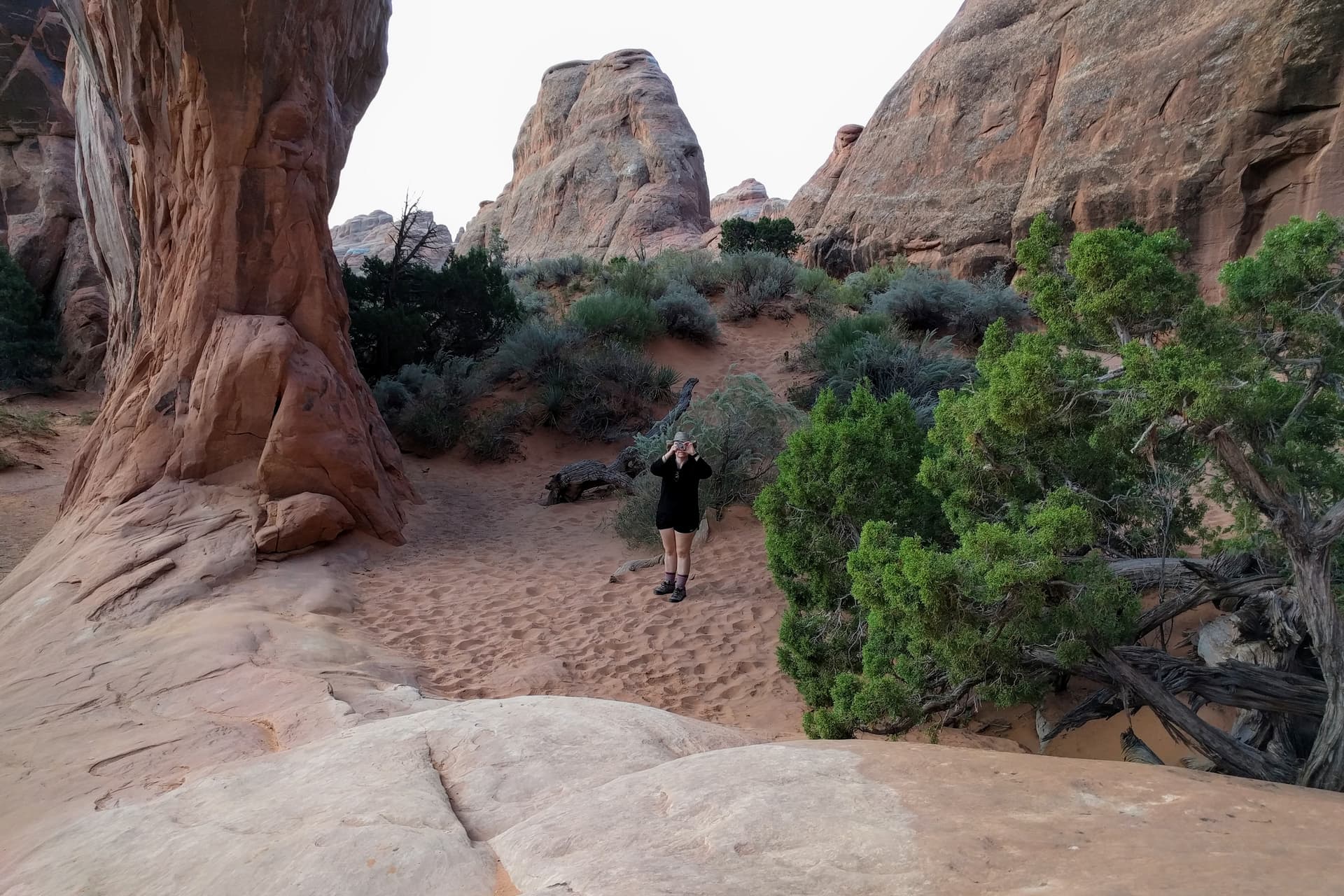 Len taking a picture of me taking a picture of her. She stands at the base of a shrub-covered dune on one side of the Pine Tree Arch, while I stand next to a Juniper tree near the middle of the arch.