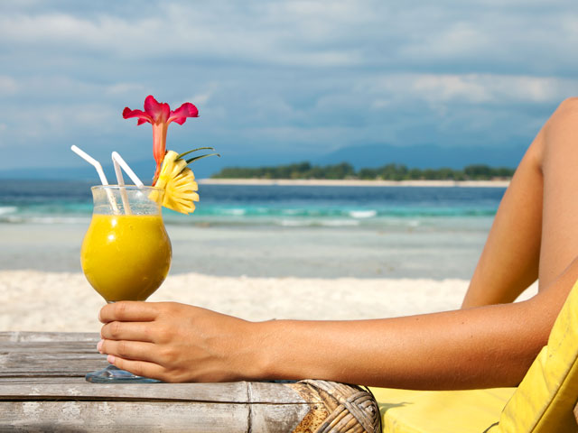 A woman is laying on a beach chair while drinking a yellow tropical drink with a pineapple garnish next to the ocean.