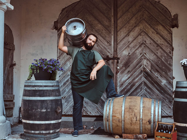 A brewer holding a keg with one foot on a beer barrel