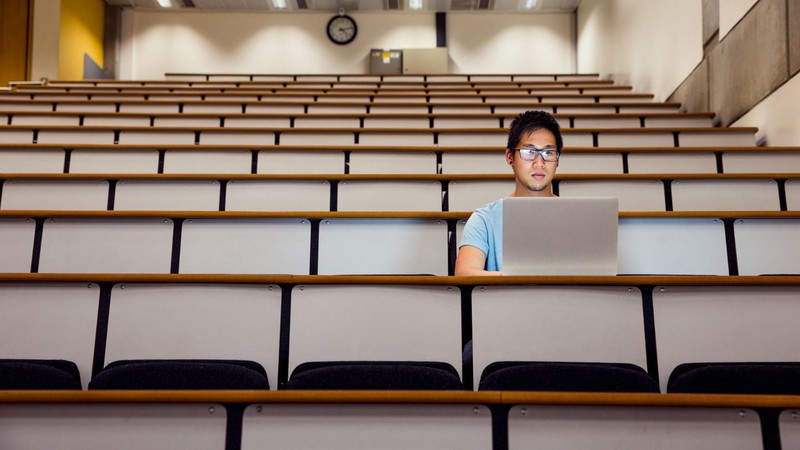 A man with glasses sits in a lecture hall while working on his computer.
