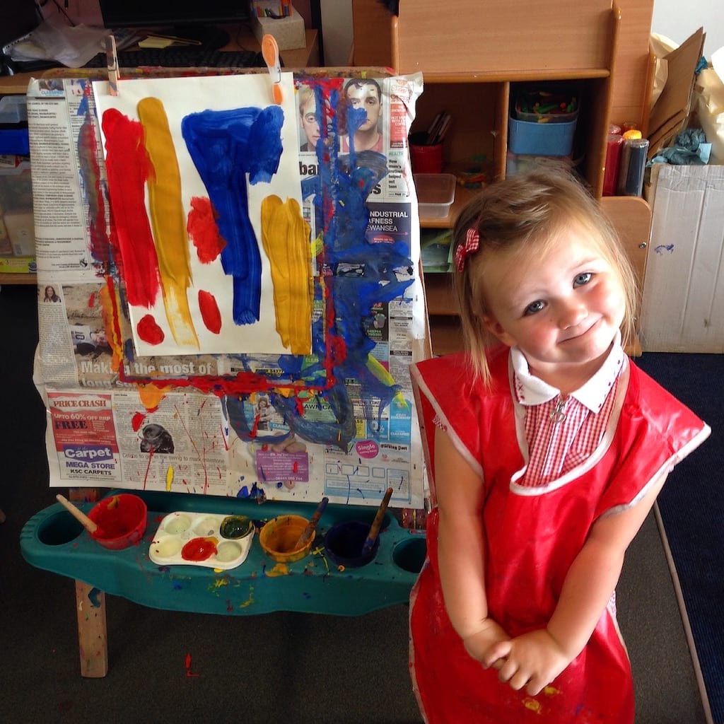 A young girl stands proudly next to an easel with a messy painting on it