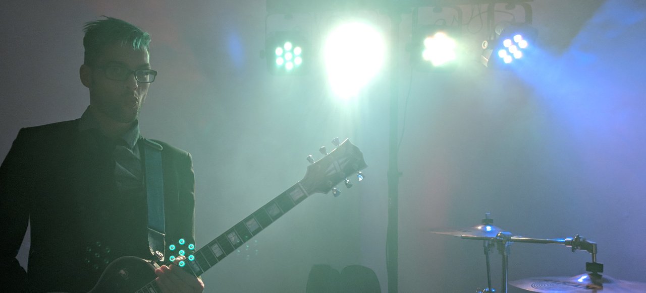 A bespectacled guitarist stood underneath some bright stage lights