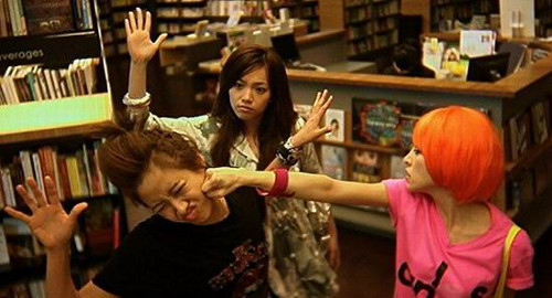 A screenshot of 3 girls fighting together in a library. A girl with bright orange hair is punching a girl in a black t-shirt. From the film 'Candy Rain'.