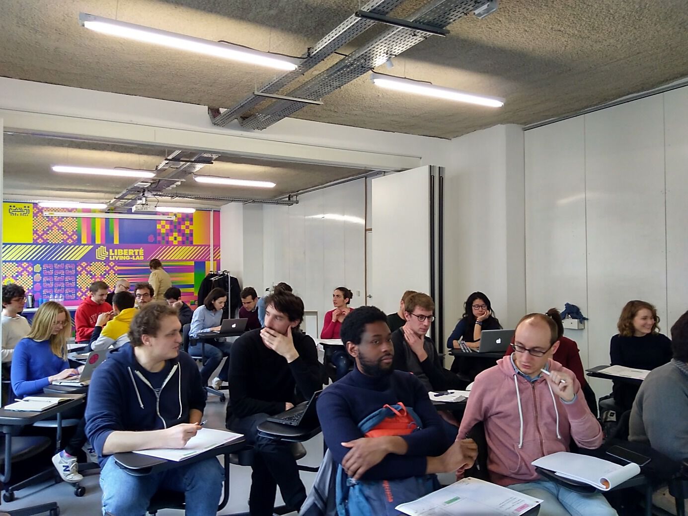 Dans une grande salle, les EIG de la promotion 2 sont assis à des pupitres individuels et discutent entre eux.