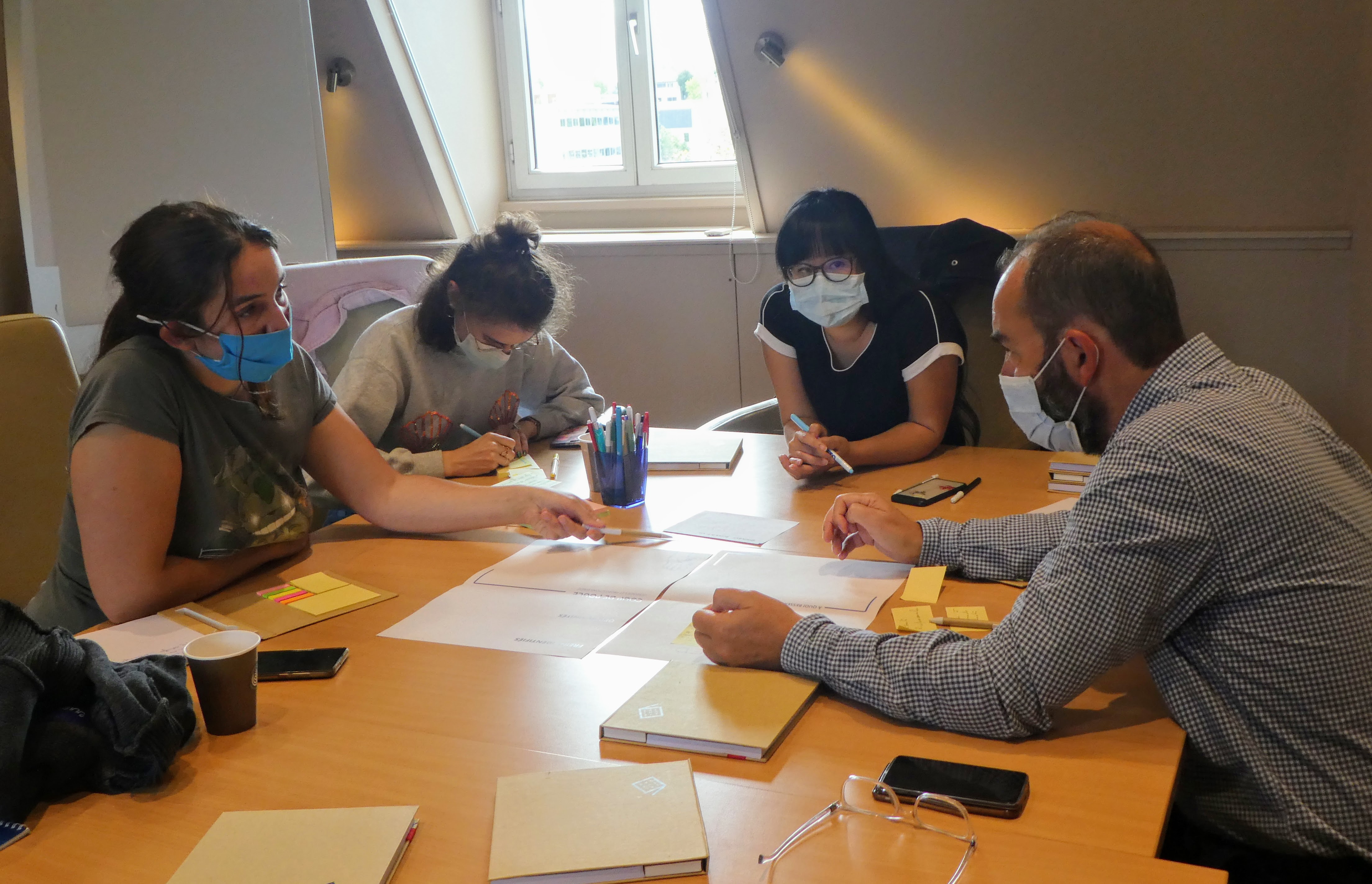 Quatre personnes sont réunies autour d'une table. Une personne écrit sur un post-it. Trois personnes sont engagés dans une discussion