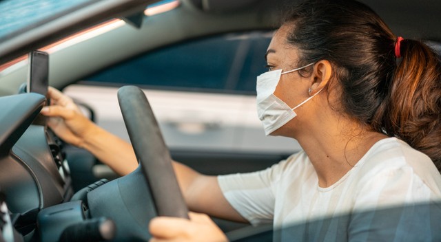 A woman wearing a mask in the driver&#039;s seat looking at a cell phone.