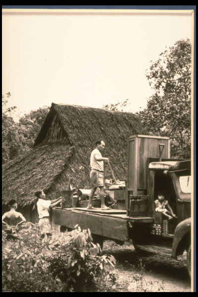 A group of individuals work to load their furniture onto a lorry in front of a thatched kampong house.