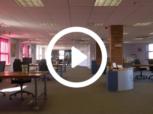 Spacious ceiling tiled office workspace containing multiple wooden desks and black office chairs next to red brick pillars Microsoft Teams background