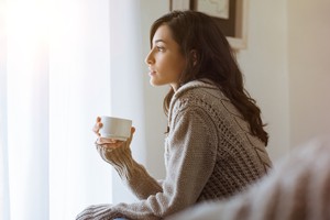 Contraception myth Young woman looking over window pane holding coffee. Thoughtful woman thinking and looking away while drinking hot tea. Woman in warm sweater looking outside window while drinking tea at morning.