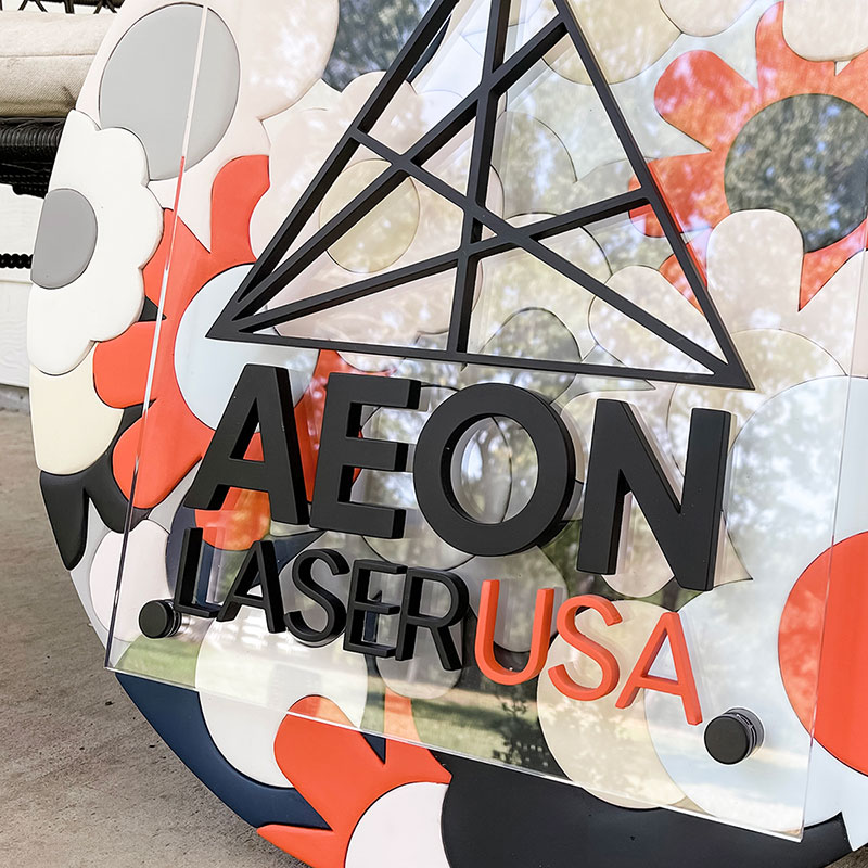 round wooden sign made of orange, blue, and white flowers with a prism and Aeon Laser USA logo on top