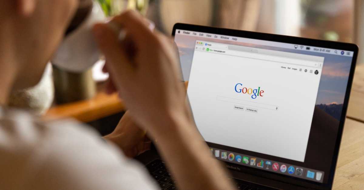 Blick über die Schulter einer Person, die vor einem Laptop sitzt und aus einer Tasse trinkt. Auf dem Laptop ist die Website Google.de zu sehen