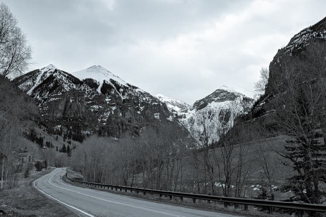 A black-and-white photo of a mountain road winding along a valley next to a tree-lined creek. Beyond the trees, three distinctly triangular snow-capped peaks can be seen.