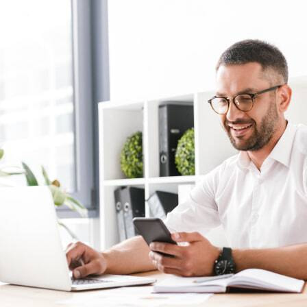 A bearded man with glasses has one hand on his laptop but is looking and smiling at his smartphone.