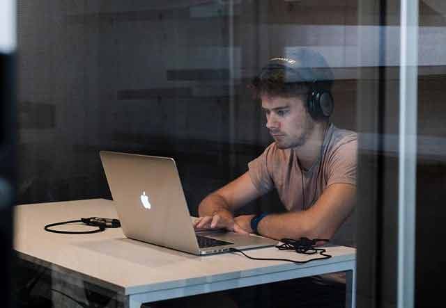 A man wearing a light brown t-shirt and black headphones working intently on his laptop