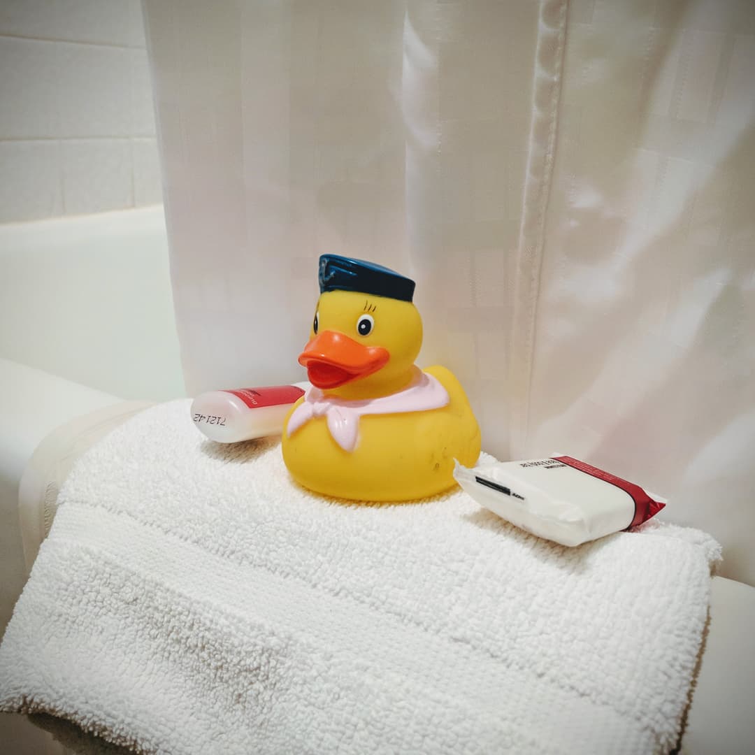 A rubber ducky perched on the rim of a hotel bathtub. To its right, a small bottle of complementary hotel shampoo, and on its left is a small bar of complimentary soap.