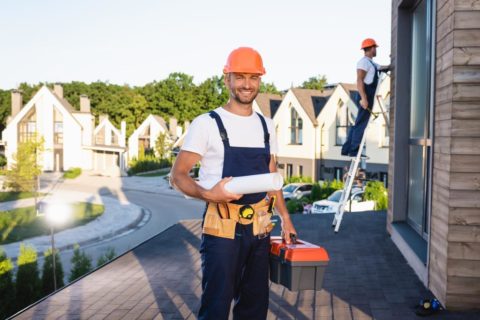 handyman with toolbox and blueprint looking at camera while colleague working on roof