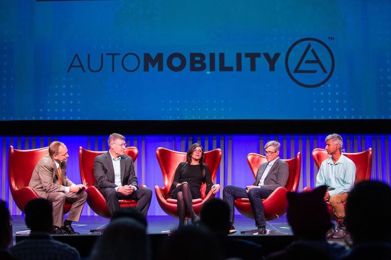 Five caucasian persons sitting on the stage of the Automotive Mobility summit in LA.