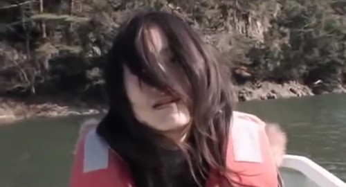 A close-up screenshot of a woman in a trance wearing a life jacket on a canoe on a lake. From the horror movie 'Noroi: The Curse'.