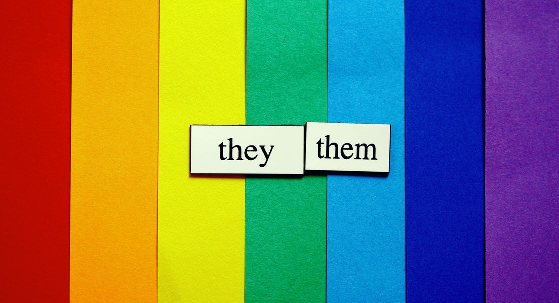 Rainbow stripes (red, orange, yellow, green, cyan, blue, purple) in the background. Pronouns “they/them” prominently placed black on white in the center.
