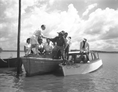 A black and white photo of leader Dato Paglar stepping from a half cabin boat into a slightly larger open boat that has been moored to a pole in the waters of Punggol. A Caucasian man helps him into the second boat, which also holds a group of onlookers.