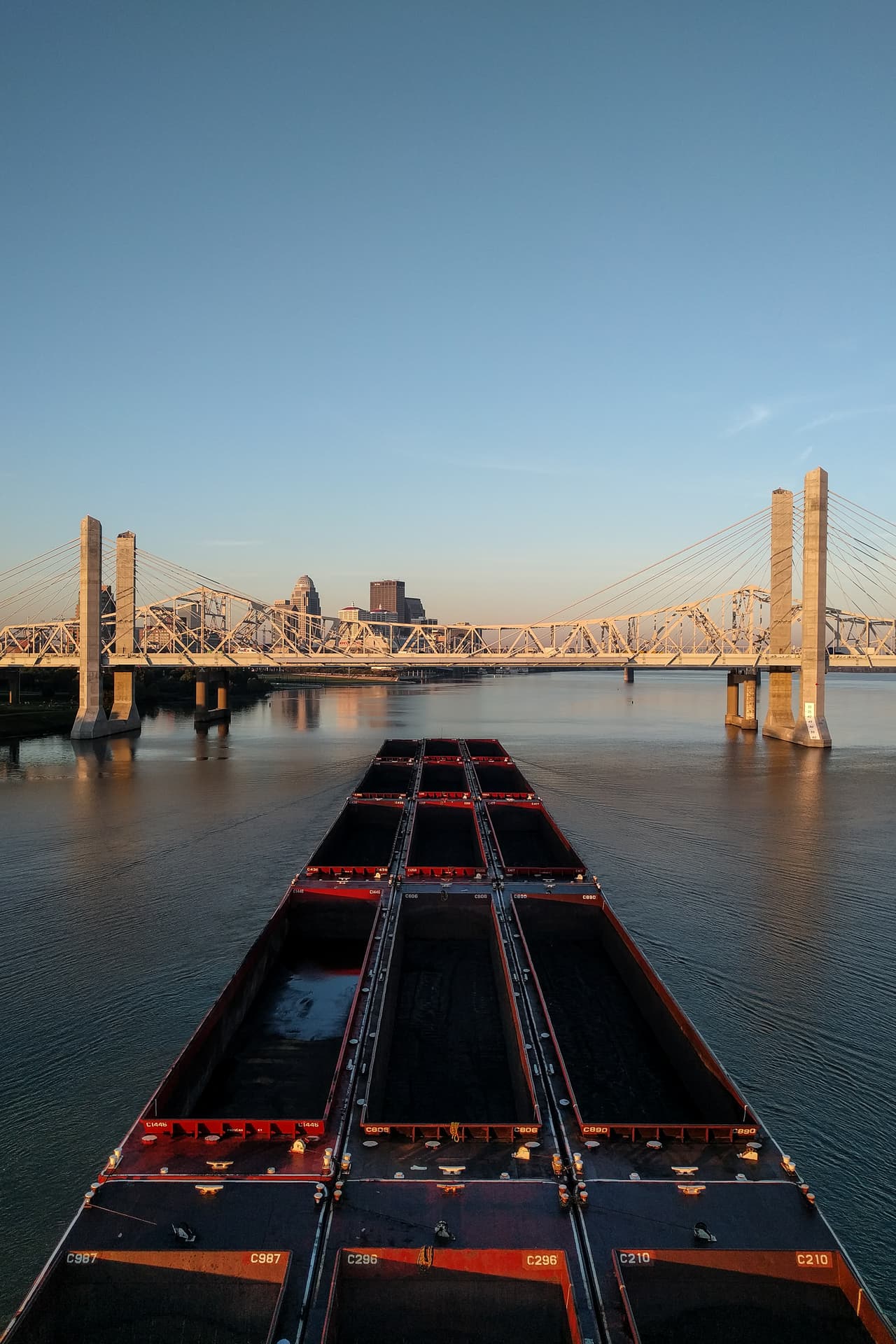 An empty coal barge approaches downtown Louisville on the Ohio River. In front of it, a brutalist suspension bridge of white steel and concrete pylons.