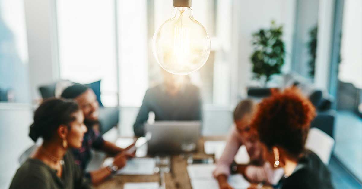 The image shows a lightbulb in the foreground, with a diverse team of people out of focus behind it. Learn how to foster creativity in your team.