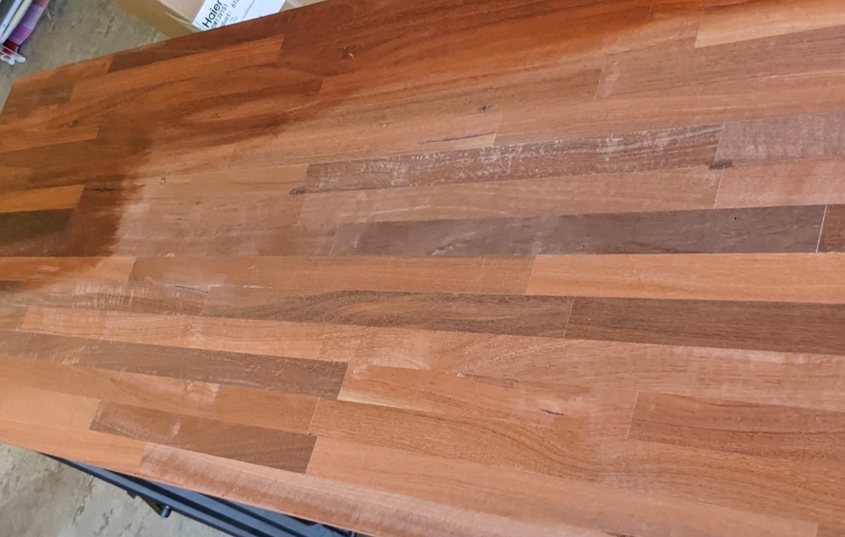 A Jarrah wooden desk top that is partially oiled, the oiled portion is a much richer and darker colour than the bare wood