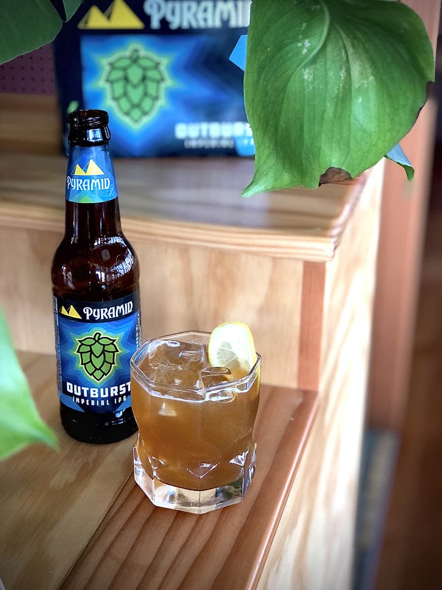 Best simple recipe summer beer cocktail for hot weather A tumbler full to the brim of the Outburst IPA beer cocktail called the Pacific Summer. The beer, peach brandy, and tea cocktail is positioned next to a bottle of Outburst IPA and some greenery.