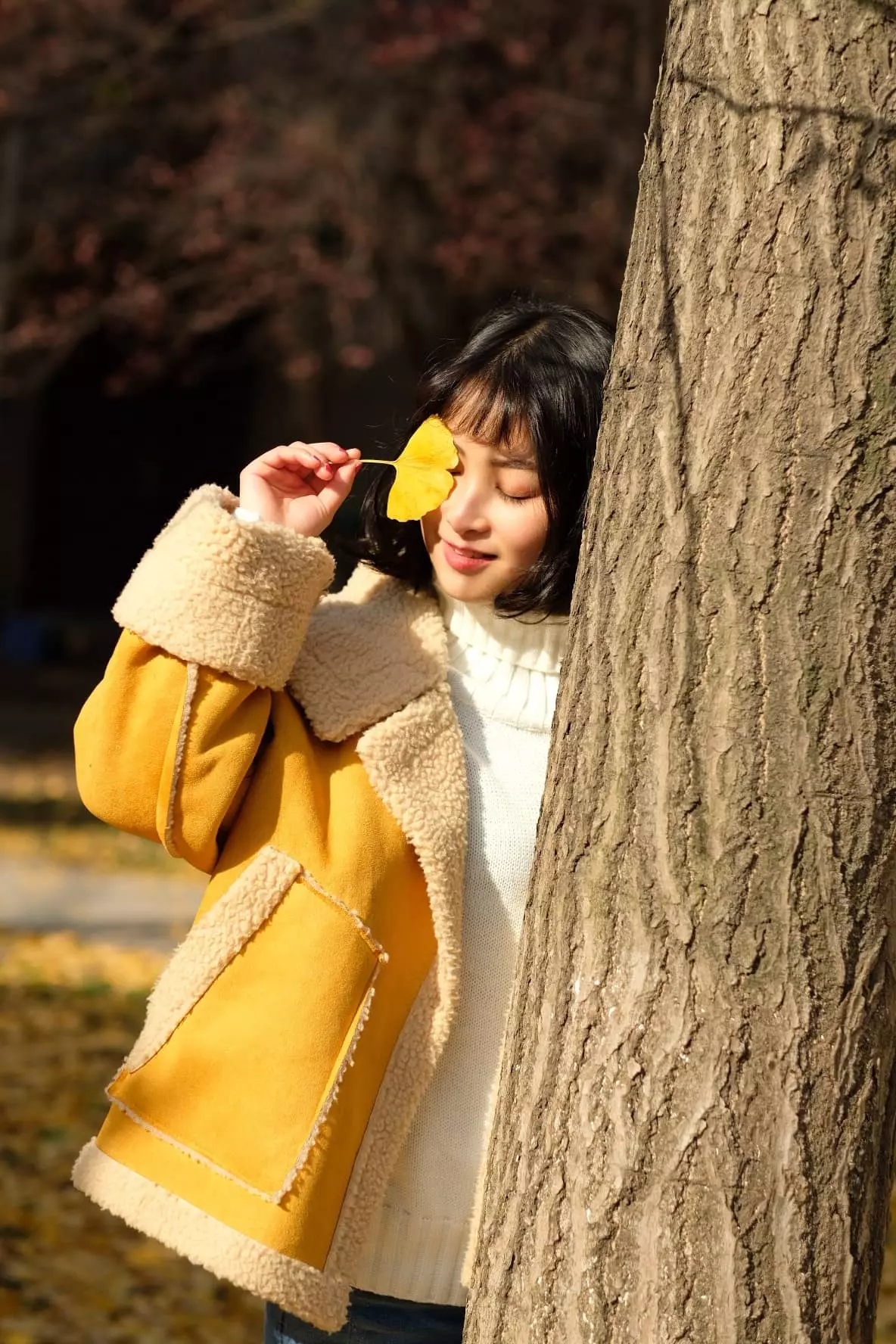 woman in yellow holding a flower to her right eye while slightly leaning on a tree