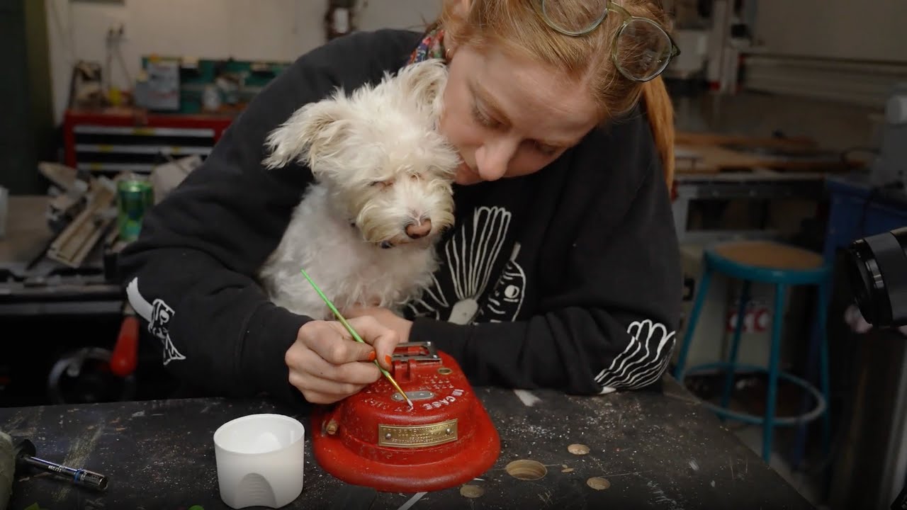 A woman sits at a desk, painting the letters of an old fire alarm. There's a dog in her lap following the paintbrush's motions.