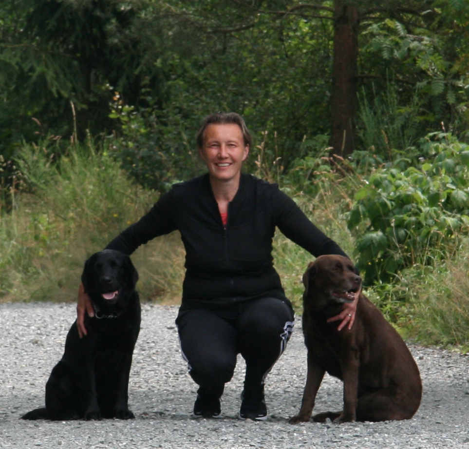 Linda met een zwarte en bruine labrador in een bosachtige omgeving.